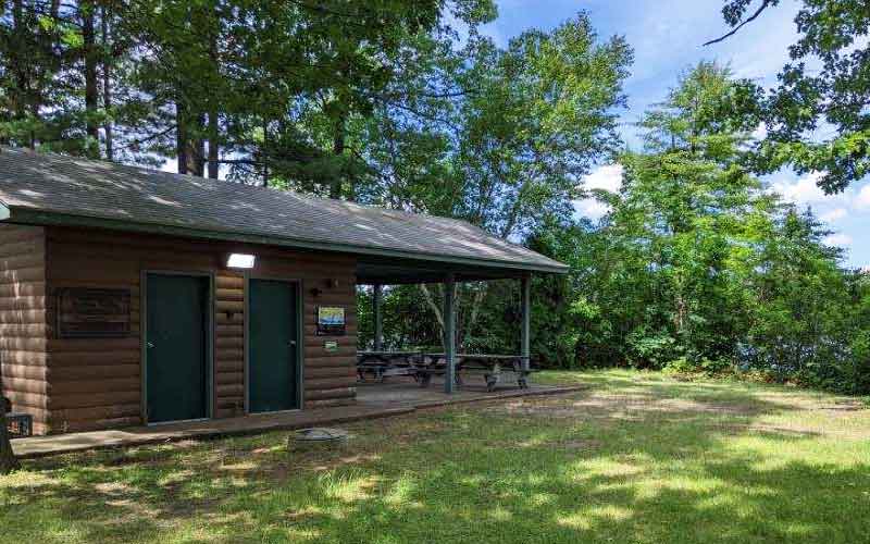 Small, brown log cabin with two doors and outdoor patio, surrounded by grass and trees