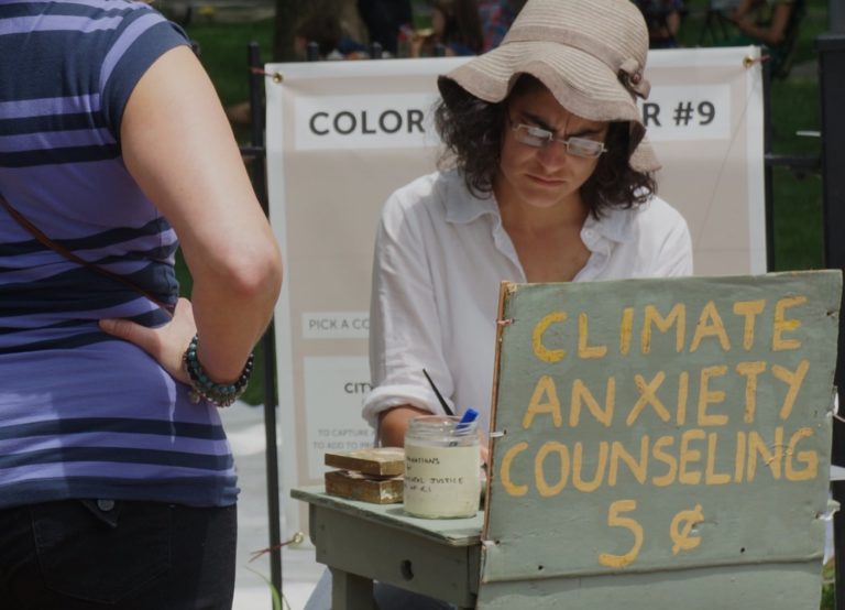 Kate Schapira, siting at a table outside with a sign that says "Climate Anxiety Counseling 5 cents"