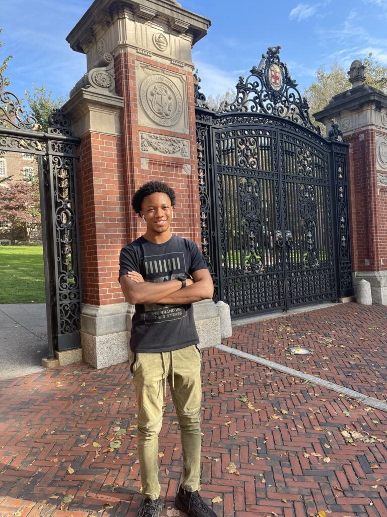A young, Black man smiling with arms crossed standing in front of a tall gate at Brown University. He is wearing a black tee shirt and beige pants