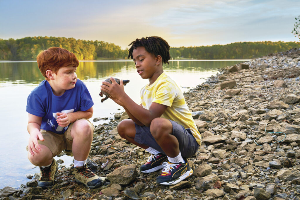 Two children squatting near a rocky shoreline, one holding a turtle.