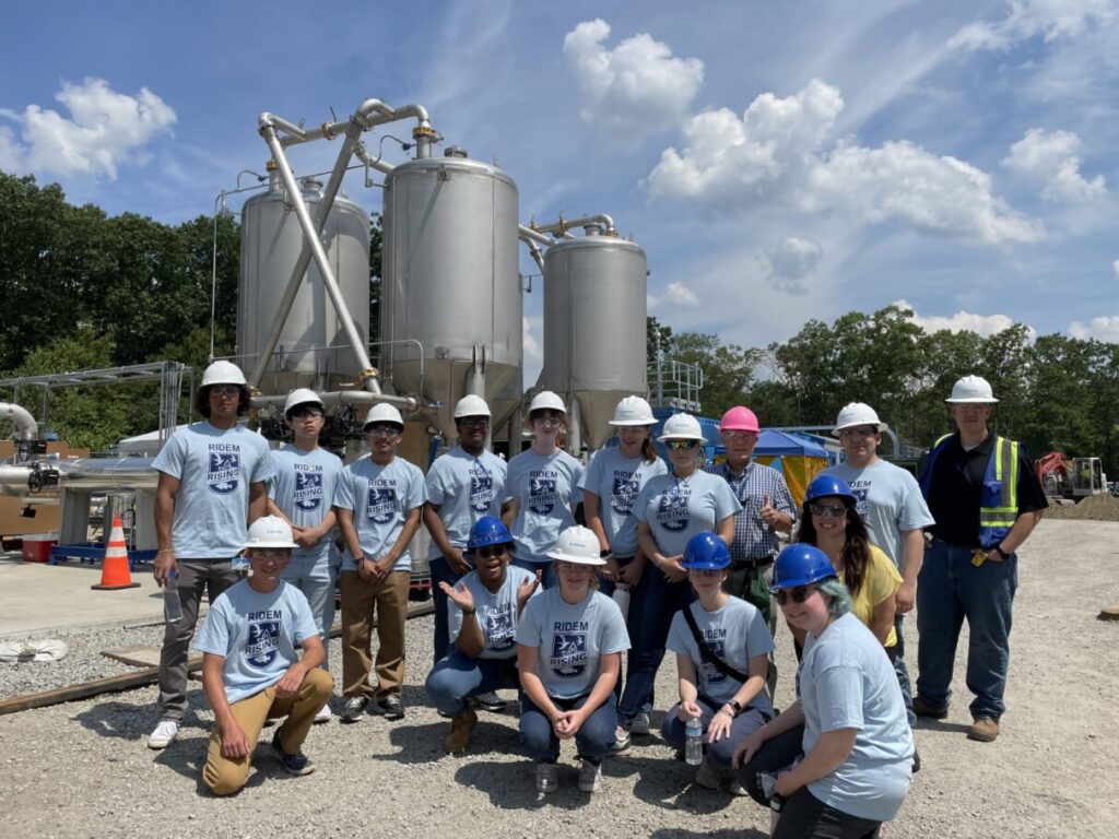 Group of young people wearing hard hats posing in front of large tanks