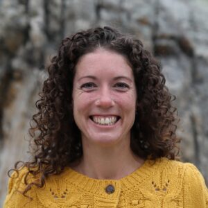 Image of Beth Alaimo, a RIEEA board member. She is grinning at the camera and her face and shoulders are in clear focus, while the background is naturey but blurred. Her hair is brown and curley and she has a nose stud on the left side of her face. Her sweater is knit and yellow.