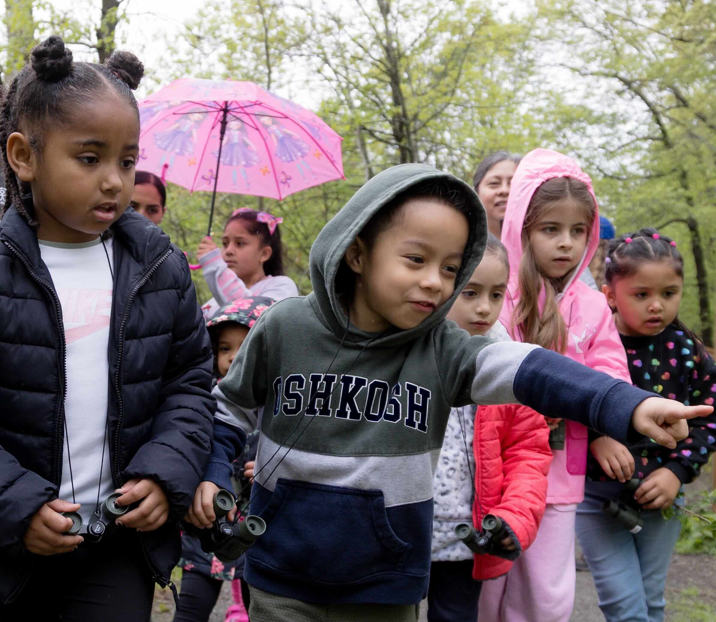 Group of young children wearing raincoats and holding umbrellas, one pointing, and all looking excited